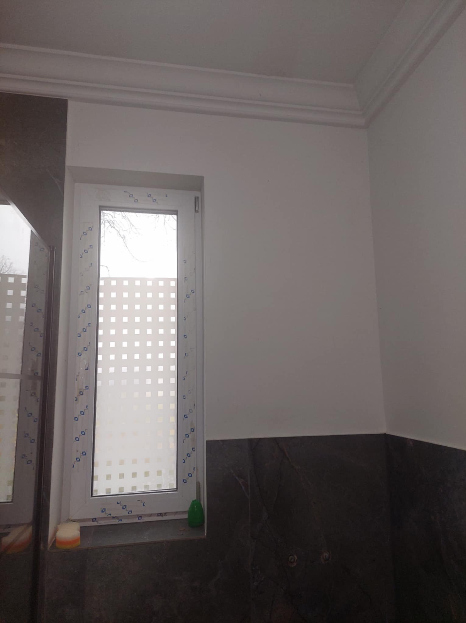 Wykończona łazienka z białymi ścianami, oknem z matową folią w kratkę i ciemnymi płytkami imitującymi marmur na dole ściany.