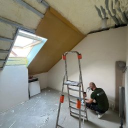 THERMAT Mateusz Dzierżęga - Prace wykończeniowe na poddaszu: ocieplenie pianką PUR, montaż stelaża, okno dachowe. Pracownik w masce aplikuje pianę. Widoczna drabina i narzędzia.