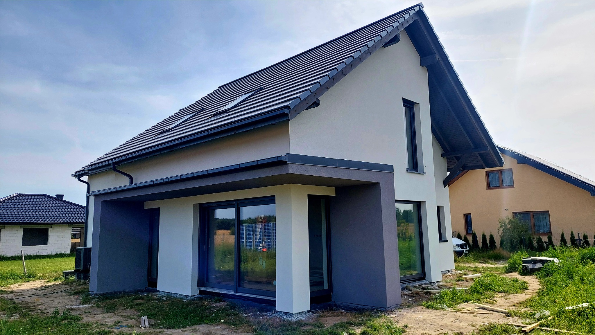 Nowoczesny dom jednorodzinny z jasną elewacją i ciemnoszarą stolarką okienną, widoczny z zewnątrz w słoneczny dzień. Minimalistyczny design, dwuspadowy dach z oknem połaciowym.