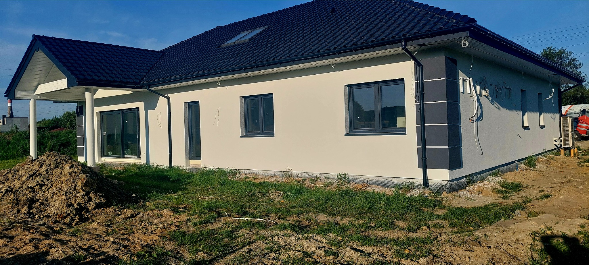 Elewacja domu jednorodzinnego po tynkowaniu, z widocznymi śladami po instalacjach. Świeżo położony tynk w jasnym odcieniu, grafitowe okna i orynnowanie. Teren wokół domu częściowo zagospodarowany.