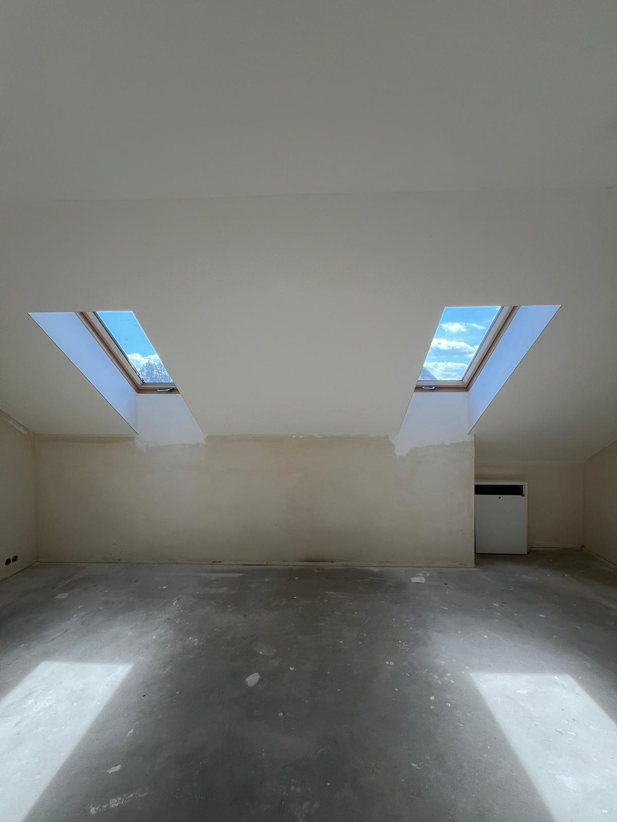 Wnętrze pokoju na poddaszu z dwoma oknami dachowymi, ściany przygotowane do malowania, surowa podłoga. Widoczne ślady szpachlowania przy oknach.