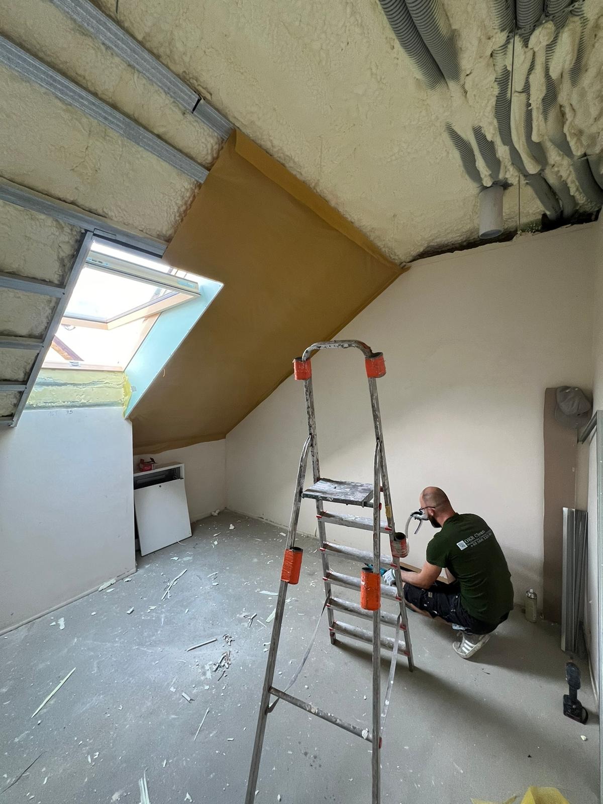 Prace wykończeniowe na poddaszu: ocieplenie pianką PUR, montaż stelaża, okno dachowe. Pracownik w masce aplikuje pianę. Widoczna drabina i narzędzia.