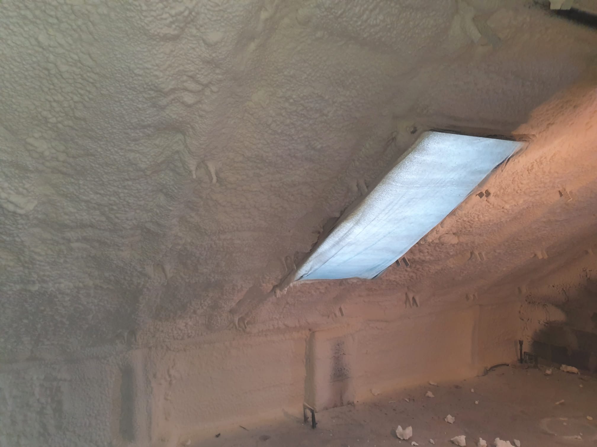 Poddasze po aplikacji pianki PUR, widoczne nierównomierne pokrycie i zabezpieczone okno dachowe. Struktura pianki przypomina gęstą, porowatą powierzchnię.