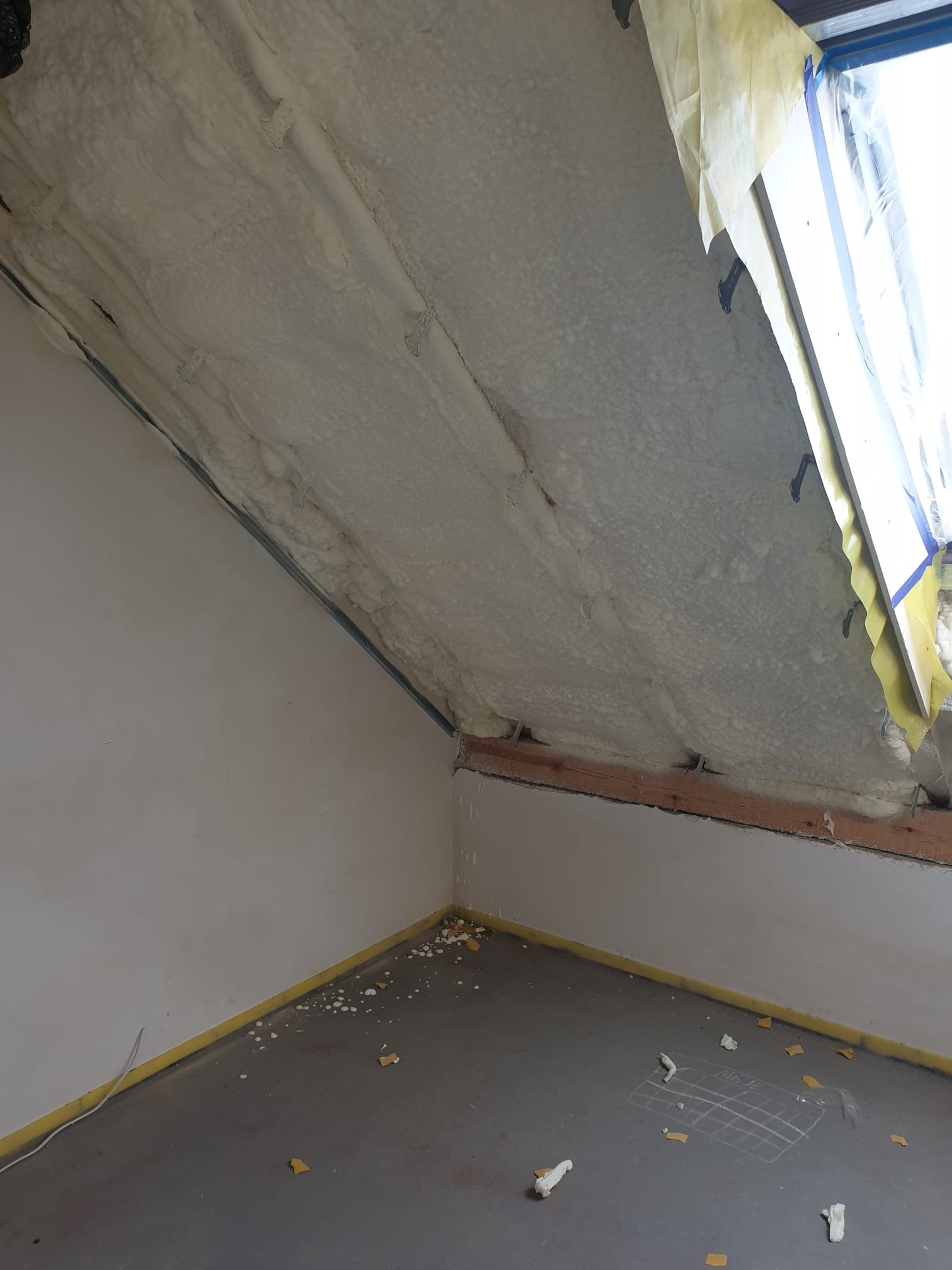 Poddasze po aplikacji pianki PUR, widoczne elementy konstrukcyjne i okno dachowe zabezpieczone folią. Na podłodze resztki pianki i szkic siatki.