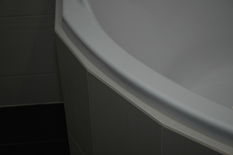 Fragment narożnej wanny obudowanej płytkami w odcieniach szarości, z widocznym fragmentem ciemnej podłogi.