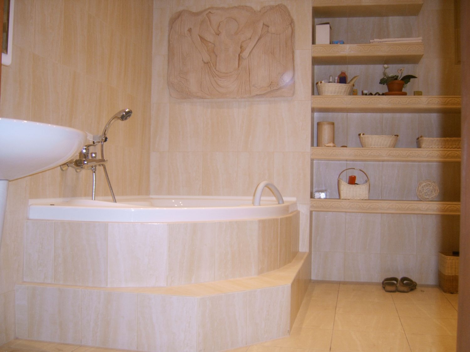 Jasna łazienka z wanną na podeście wyłożonym płytkami imitującymi marmur, umywalką na postumencie i zdobioną płaskorzeźbą na ścianie oraz półkami z wiklinowymi koszykami.