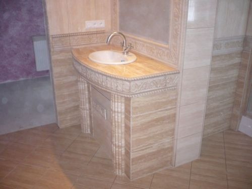 Ukończona łazienka z beżowymi płytkami imitującymi trawertyn, umywalka nablatowa z ozdobnym blatem i chromowaną baterią, widoczne gniazdko elektryczne.