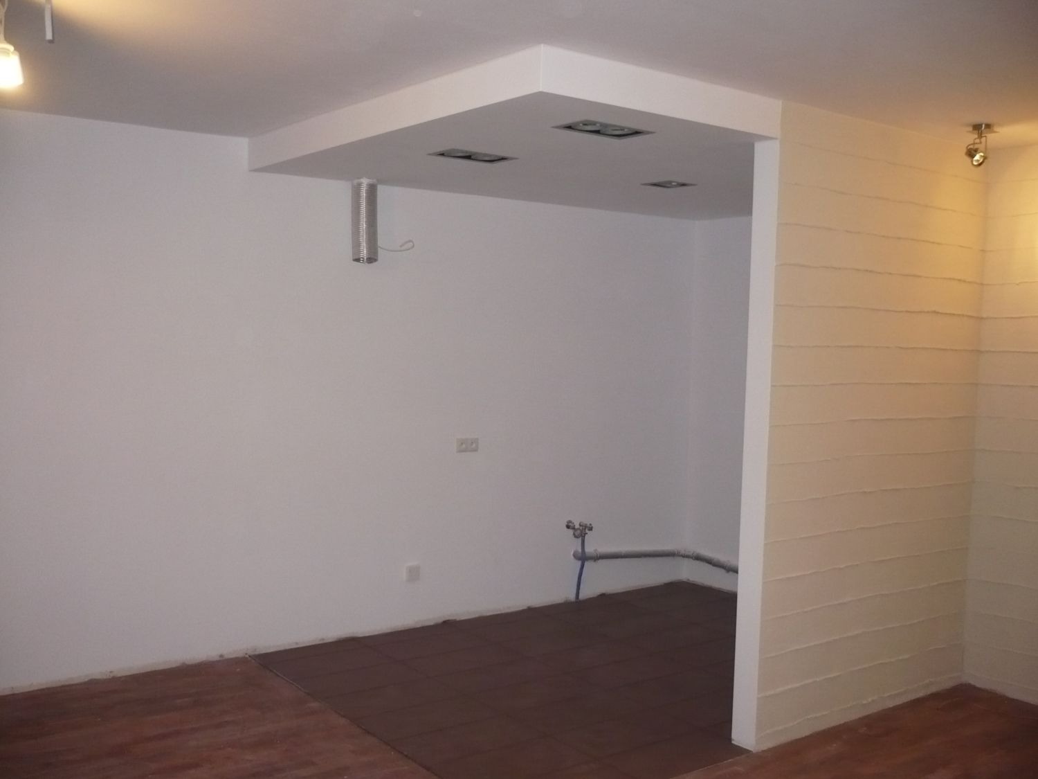 Surowe wnętrze z białymi ścianami, częściowo wykończoną podłogą z brązowych płytek i widoczną instalacją hydrauliczną przy podłodze.