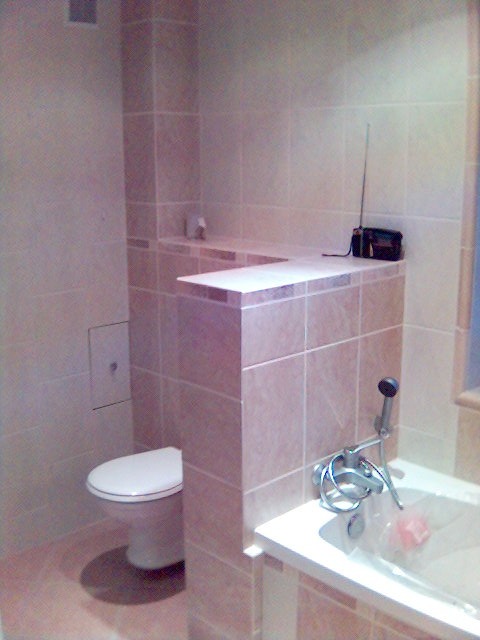 Wykończona łazienka z beżowymi płytkami, widoczna toaleta, wanna z baterią i radio na półce z płytek.