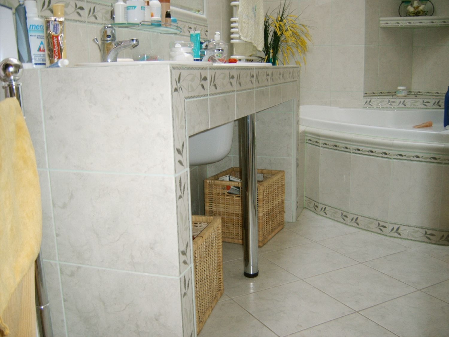 Wykończona łazienka w jasnych płytkach z motywem roślinnym, widoczna umywalka na postumencie z koszami wiklinowymi i fragment wanny.