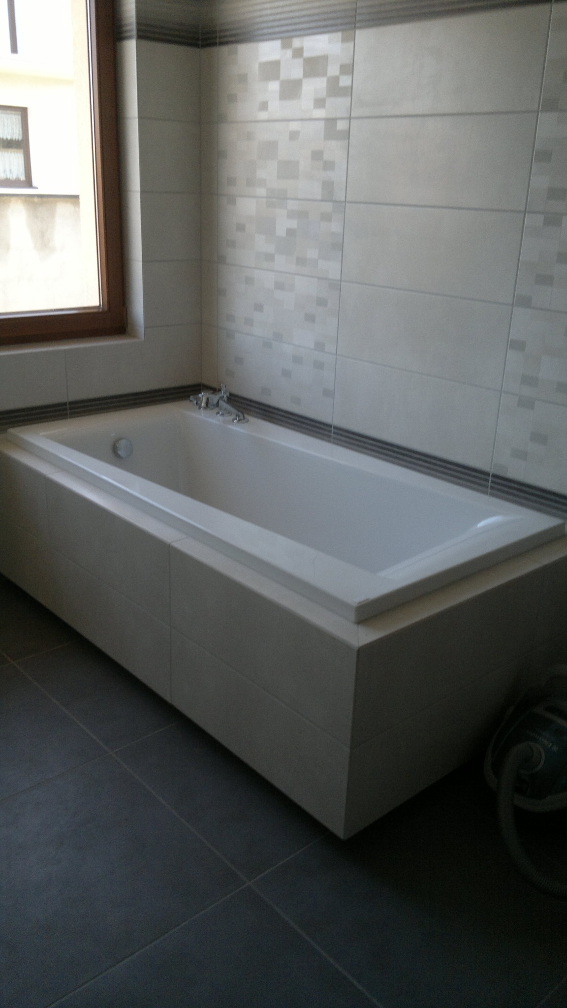Wykończona łazienka z zabudowaną wanną w obudowie z płytek, szara podłoga z płytek, ściany wyłożone jasnymi płytkami z dekoracyjnymi wstawkami, widoczne okno i odkurzacz w rogu.