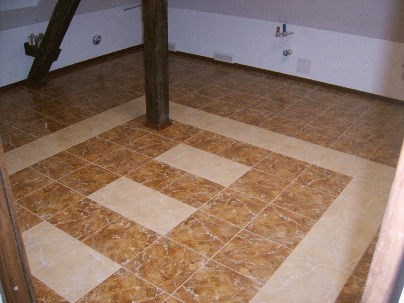 Ukończona podłoga w poddaszu z brązowych i beżowych płytek ułożonych w kwadraty, widoczny drewniany słup podtrzymujący strop i fragment instalacji hydraulicznej na ścianie.