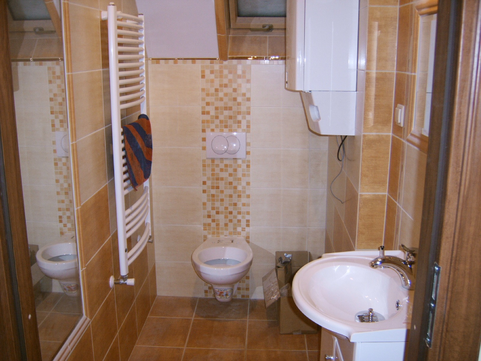 Umeblowana łazienka z beżowymi i brązowymi płytkami, wiszącą muszlą klozetową, grzejnikiem drabinkowym z ręcznikiem w paski i białą umywalką z chromowaną baterią.