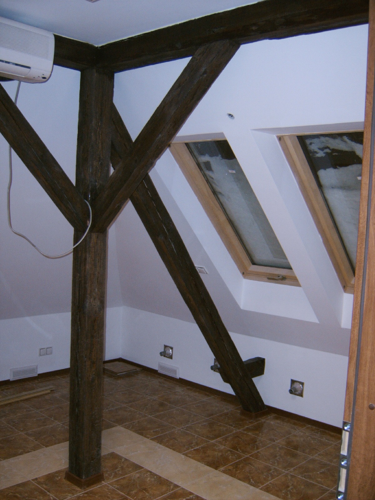 Poddasze z drewnianą konstrukcją nośną, dwoma oknami dachowymi i brązową terakotą na podłodze, widoczny klimatyzator na ścianie.
