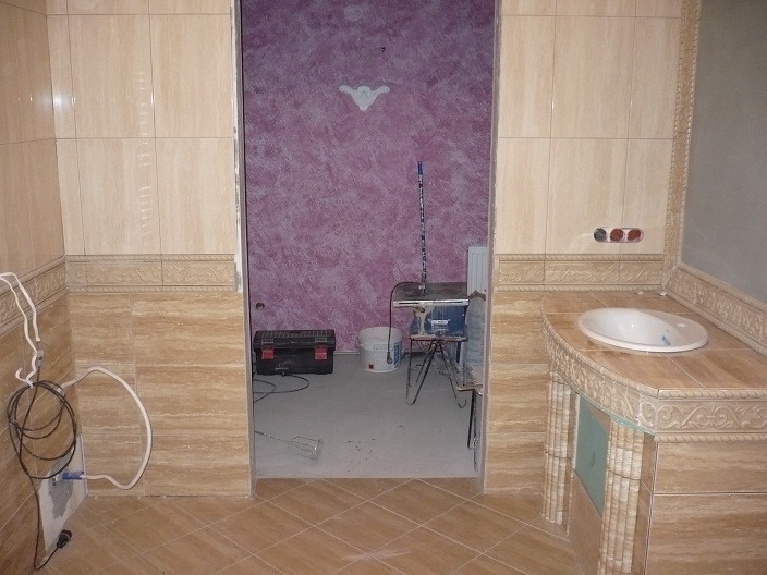 Łazienka w trakcie prac wykończeniowych: beżowe płytki, dekoracyjne listwy, umywalka z obudową, widoczne narzędzia i wiadro z farbą w tle, ściana pomalowana na fioletowo.