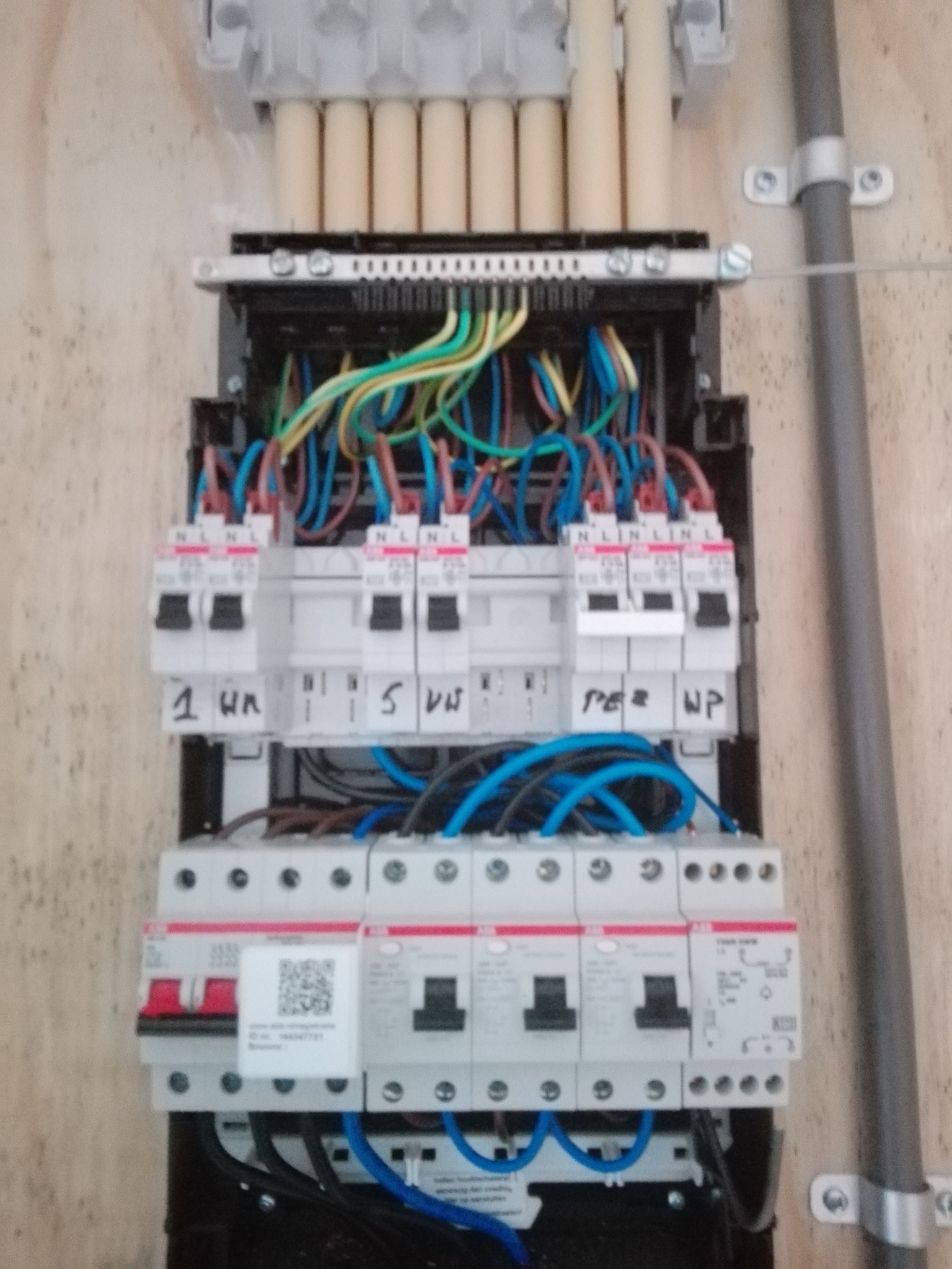 Rozdzielnica elektryczna z przewodami o różnych kolorach, zabezpieczeniami i oznaczeniami, zamontowana na ścianie.