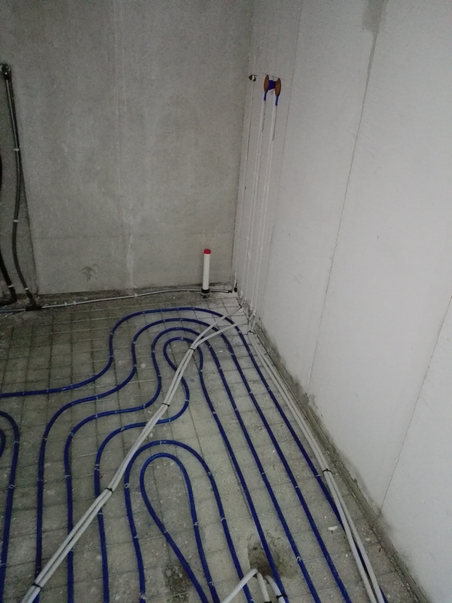 Niebieskie rury ogrzewania podłogowego ułożone na siatce w surowym wnętrzu, widoczne pionowe rury przy ścianie.