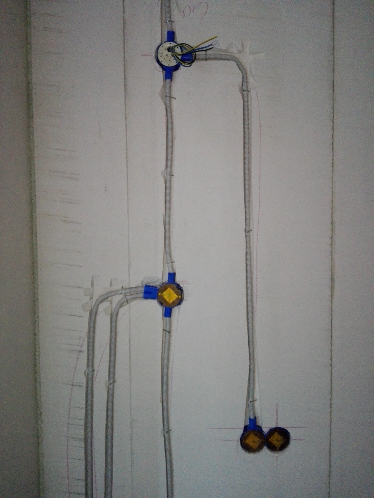 Szare rury karbowane prowadzące do puszek elektrycznych zamontowanych na surowej, białej ścianie. Widoczne kable wewnątrz puszek i ślady ołówka na ścianie.