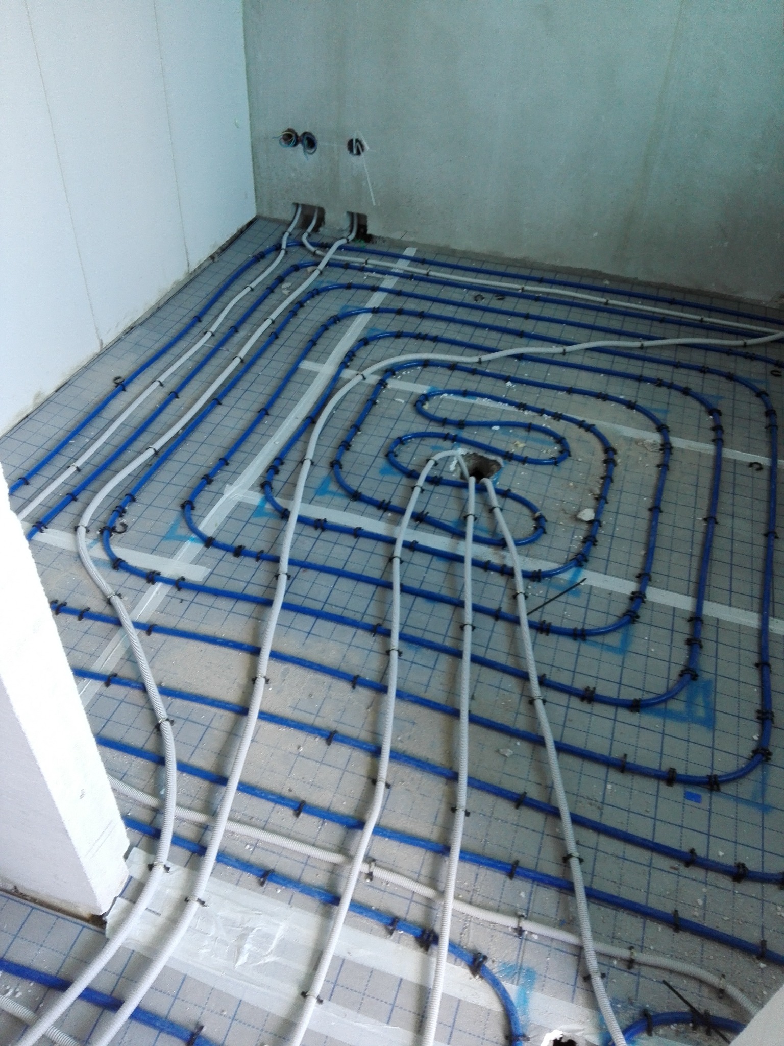 Rozłożone rury ogrzewania podłogowego, białe i niebieskie, przymocowane do podłoża siatką z niebieskimi elementami mocującymi, widoczne otwory w ścianie.