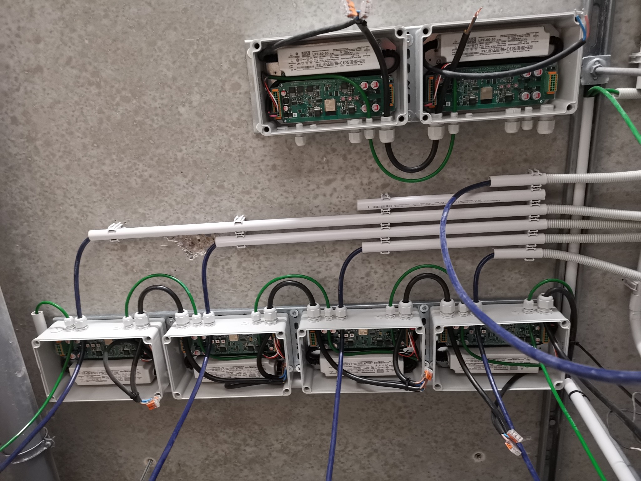 Szereg szarych puszek instalacyjnych z widocznymi sterownikami LED i okablowaniem przymocowanych do betonowego sufitu, z białymi rurkami ochronnymi na przewody.