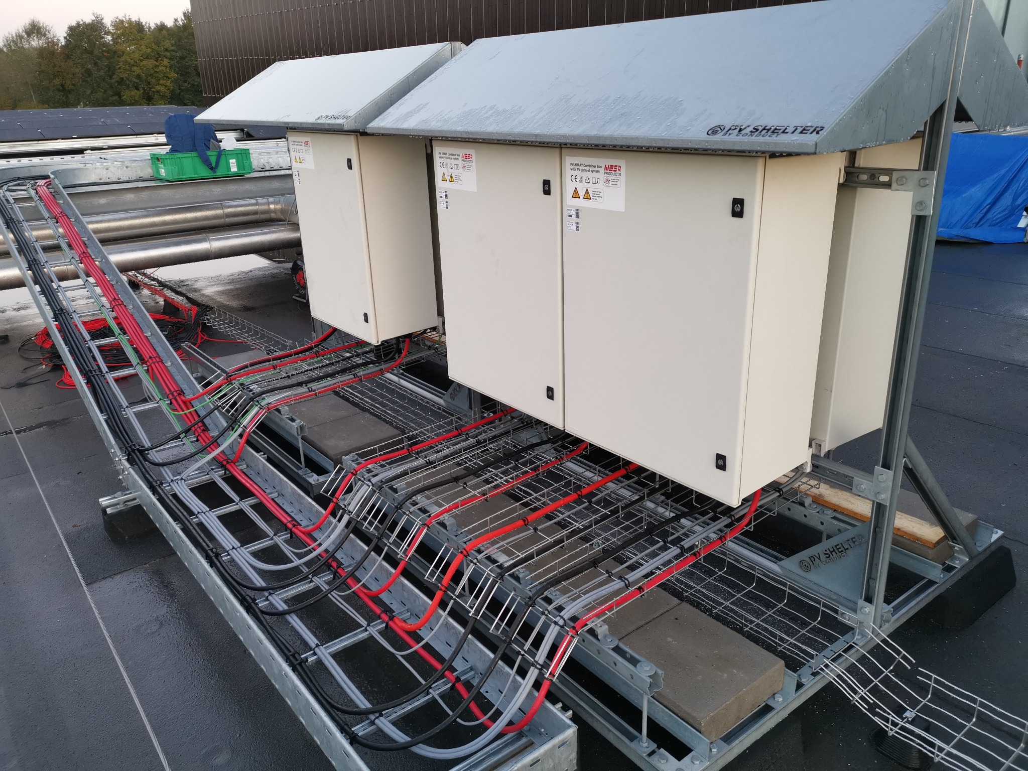 Dwie białe szafy elektryczne z szarymi daszkami, umieszczone na metalowej konstrukcji z kablami ułożonymi w korytkach na płaskim dachu.