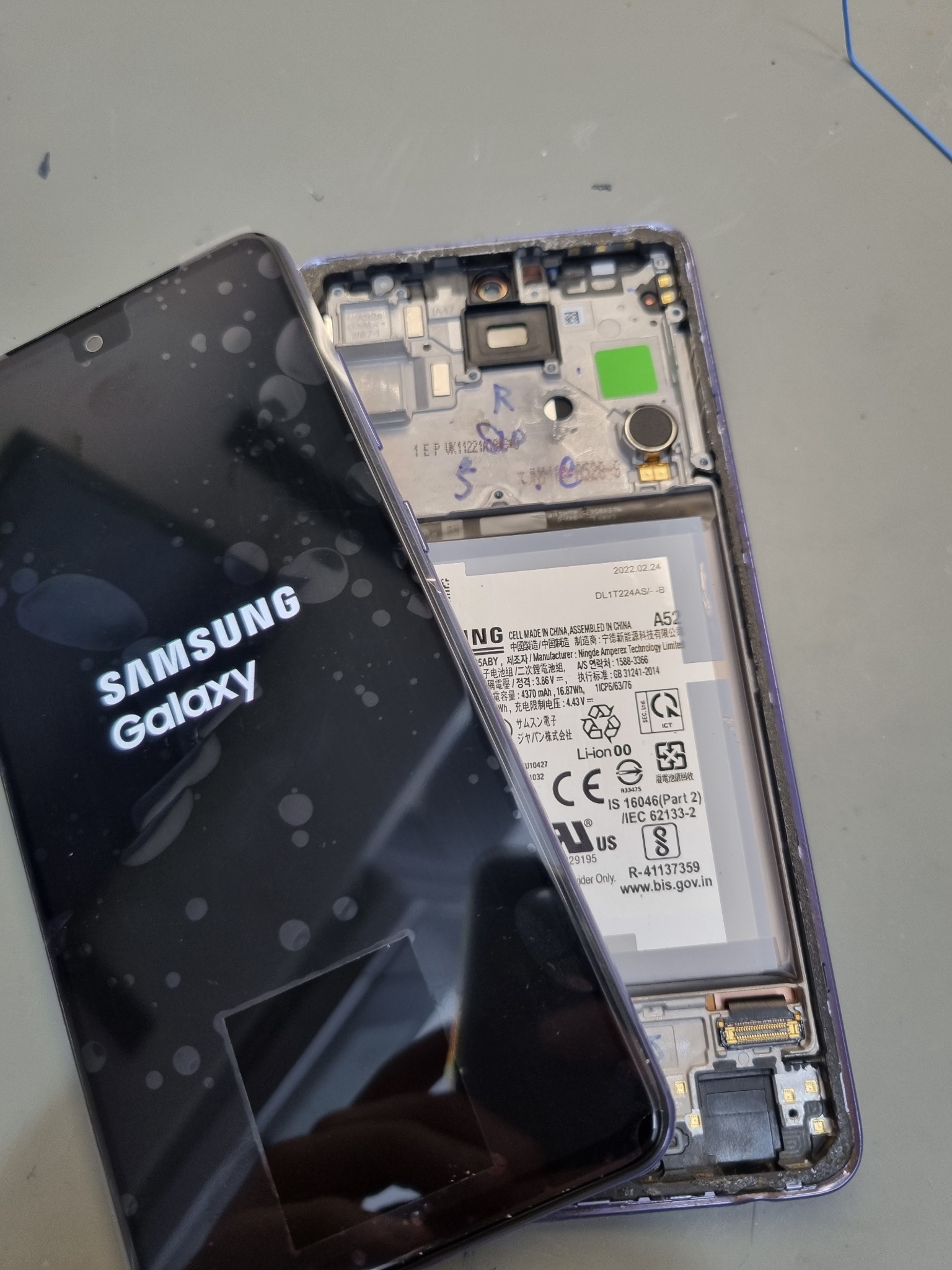 Rozłożony telefon Samsung Galaxy A52 z widocznym ekranem z logo i wnętrzem obudowy, przygotowany do wymiany ekranu, widoczne ślady po kleju na wyświetlaczu.