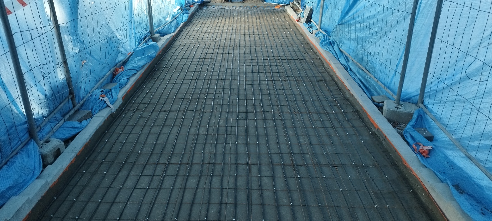 Zbrojenie pod wylewkę betonową, widoczne siatki z drutu zbrojeniowego ułożone w szalunku, zabezpieczone niebieską plandeką.