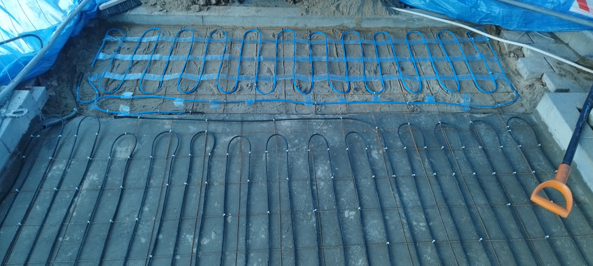 Montaż ogrzewania podłogowego: niebieskie rurki układane na siatce zbrojeniowej, widoczny fragment łopaty z pomarańczowym uchwytem.