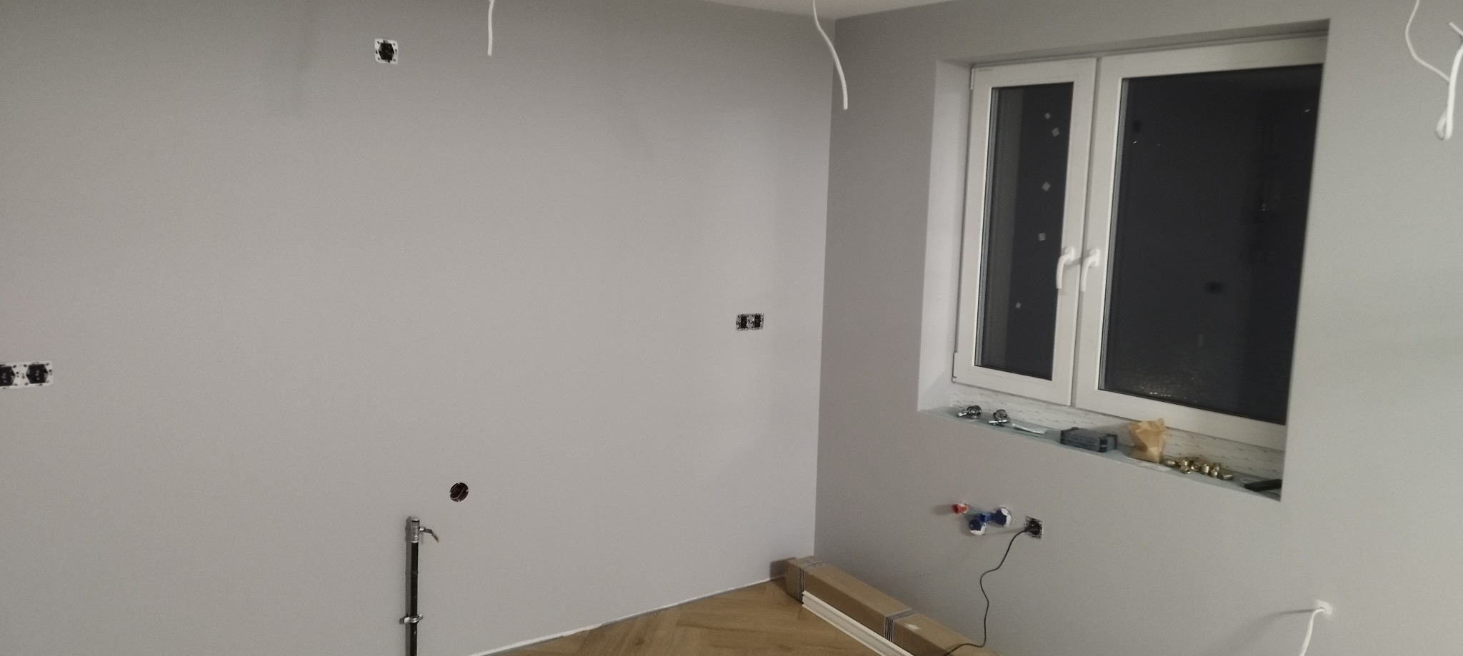 Szare wnętrze w trakcie remontu: widoczne puszki elektryczne w ścianie, wystające przewody, rury hydrauliczne i akcesoria na parapecie okna, parkiet w jodełkę na podłodze.