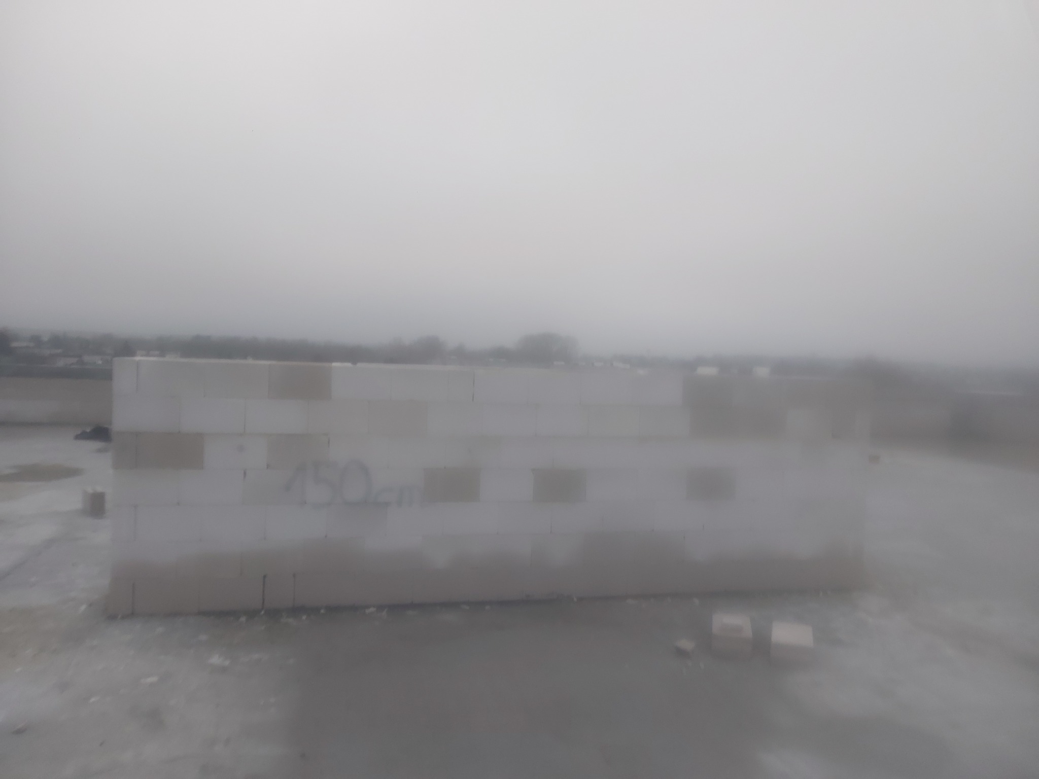 Wzniesiona ściana z białych bloczków z napisem '150cm' w szarych warunkach pogodowych na placu budowy.