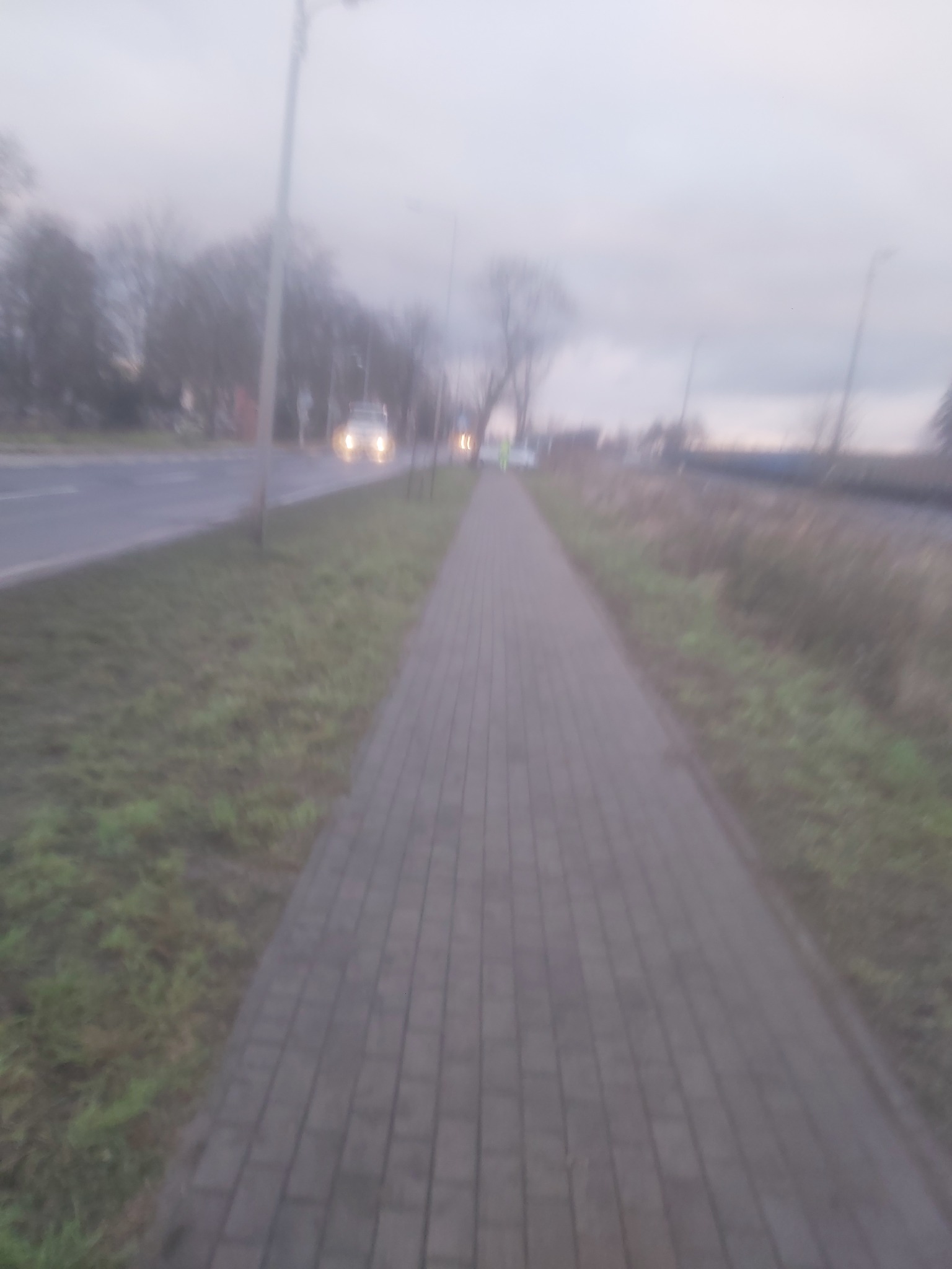 Szary chodnik z kostki brukowej wzdłuż drogi w pochmurny dzień, widoczne światła samochodów w oddali.