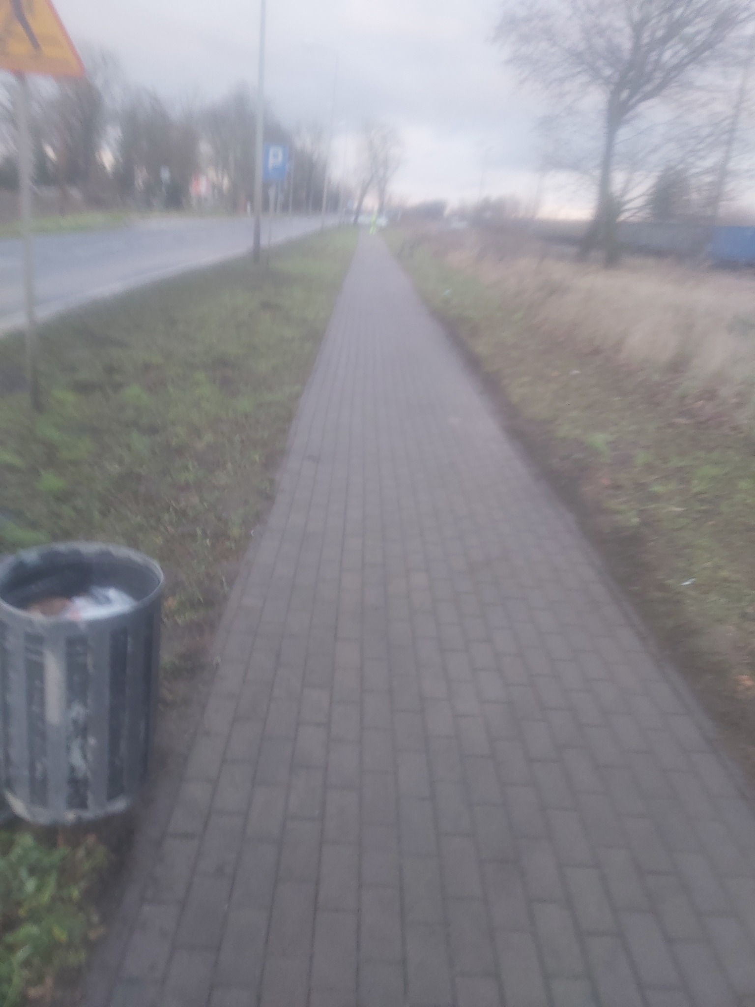 Szara kostka brukowa na chodniku wzdłuż ulicy w pochmurny dzień, z widocznym koszem na śmieci po lewej stronie i znakami drogowymi w tle.