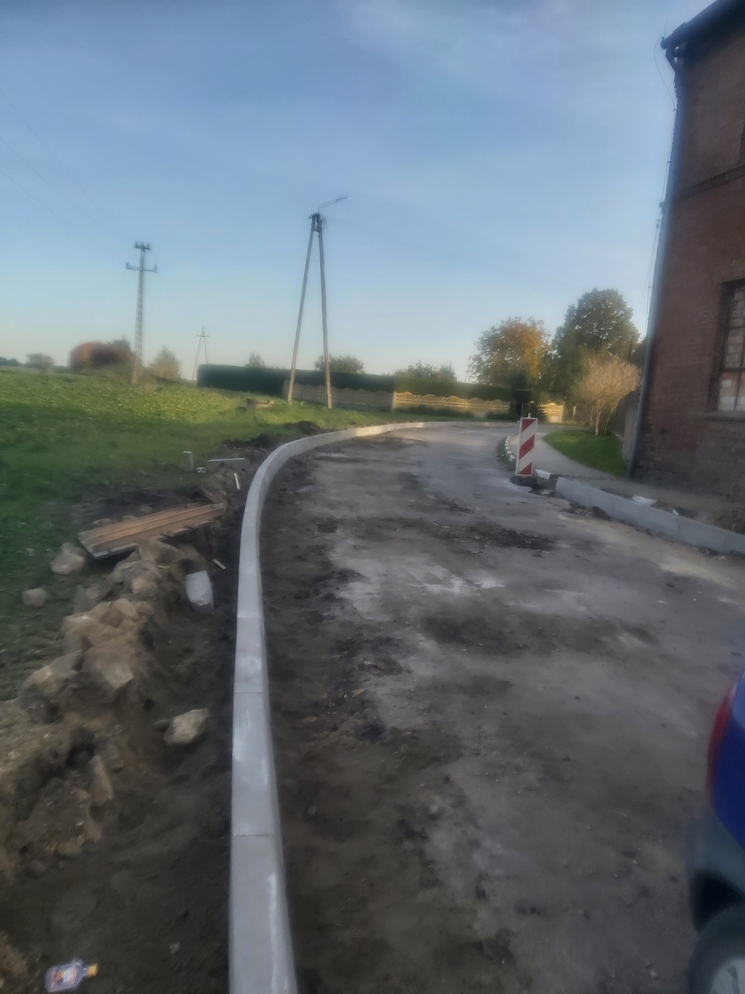Krawężniki betonowe ułożone wzdłuż drogi gruntowej, widoczne przygotowania do dalszych prac brukarskich, w tle budynek z czerwonej cegły i słupy energetyczne.