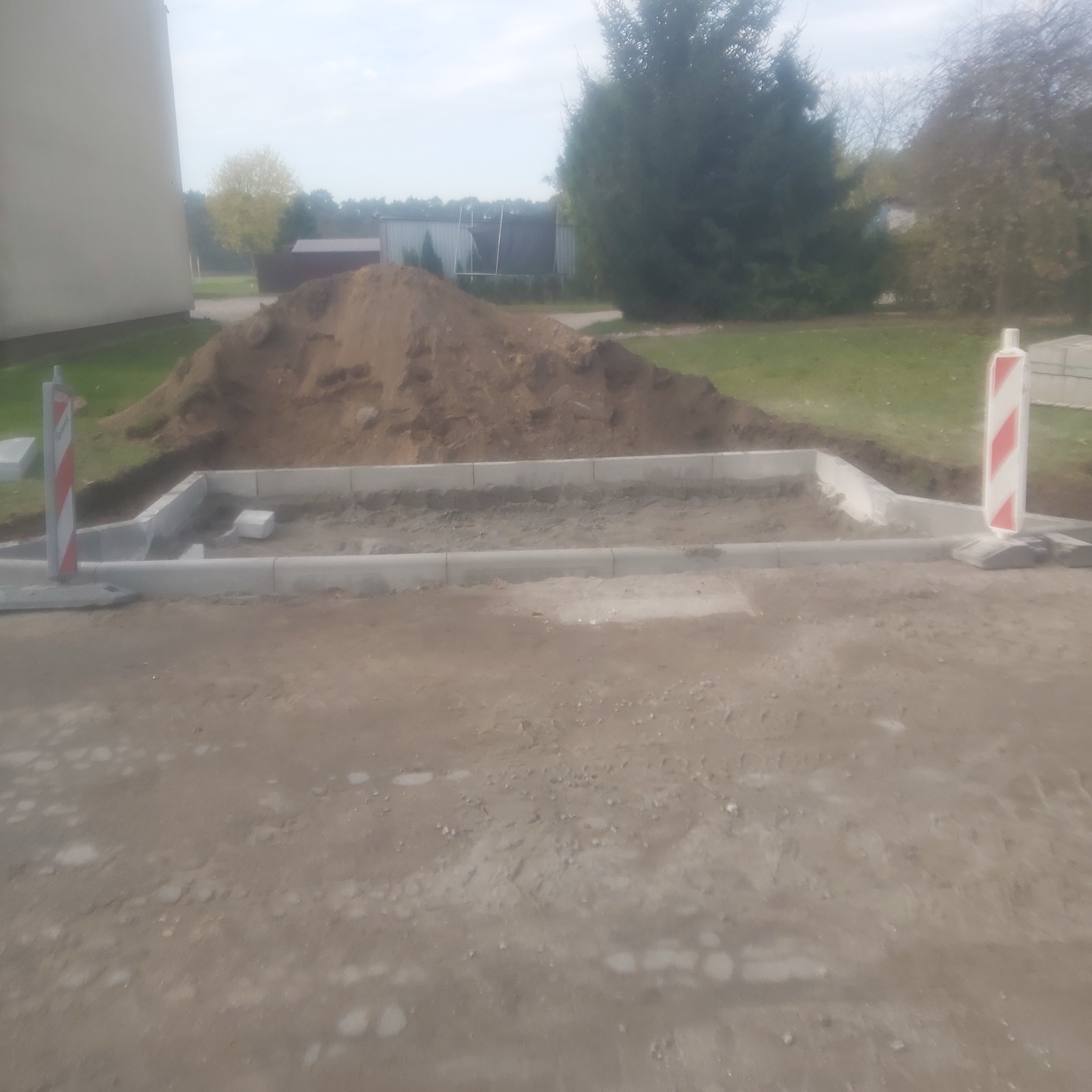 Przygotowanie podłoża pod kostkę brukową: ułożone obrzeża betonowe, stos piasku, widoczne pale ostrzegawcze.