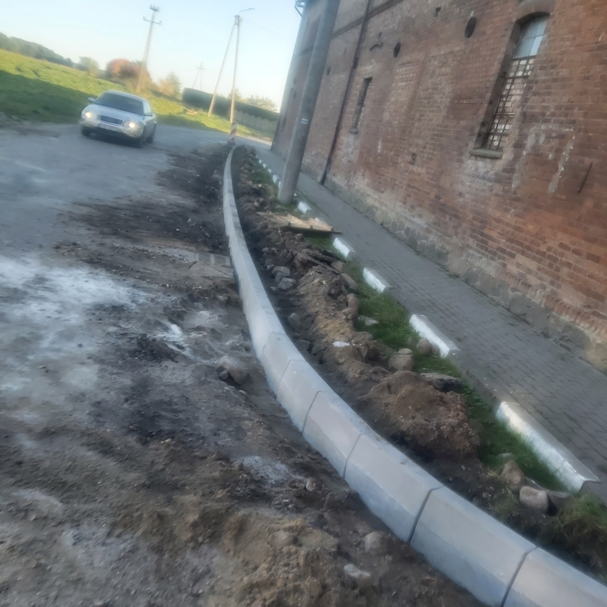 Nowo ułożone obrzeża chodnikowe z szarej kostki betonowej, oddzielające jezdnię od chodnika z czerwonej cegły, z samochodem w tle.