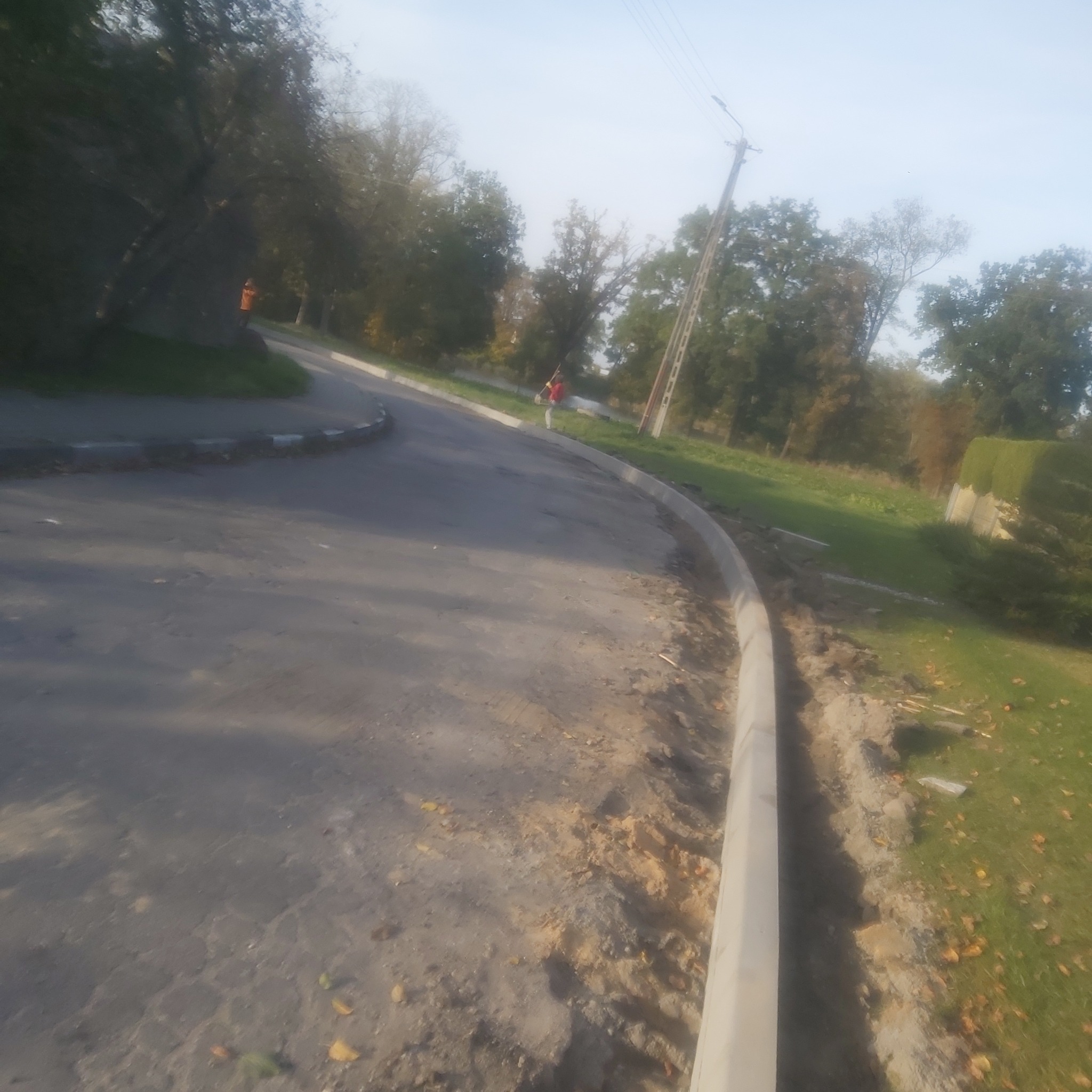 Nowo ułożony, jasnoszary krawężnik oddzielający asfaltową drogę od trawnika, z pracownikiem w oddali.