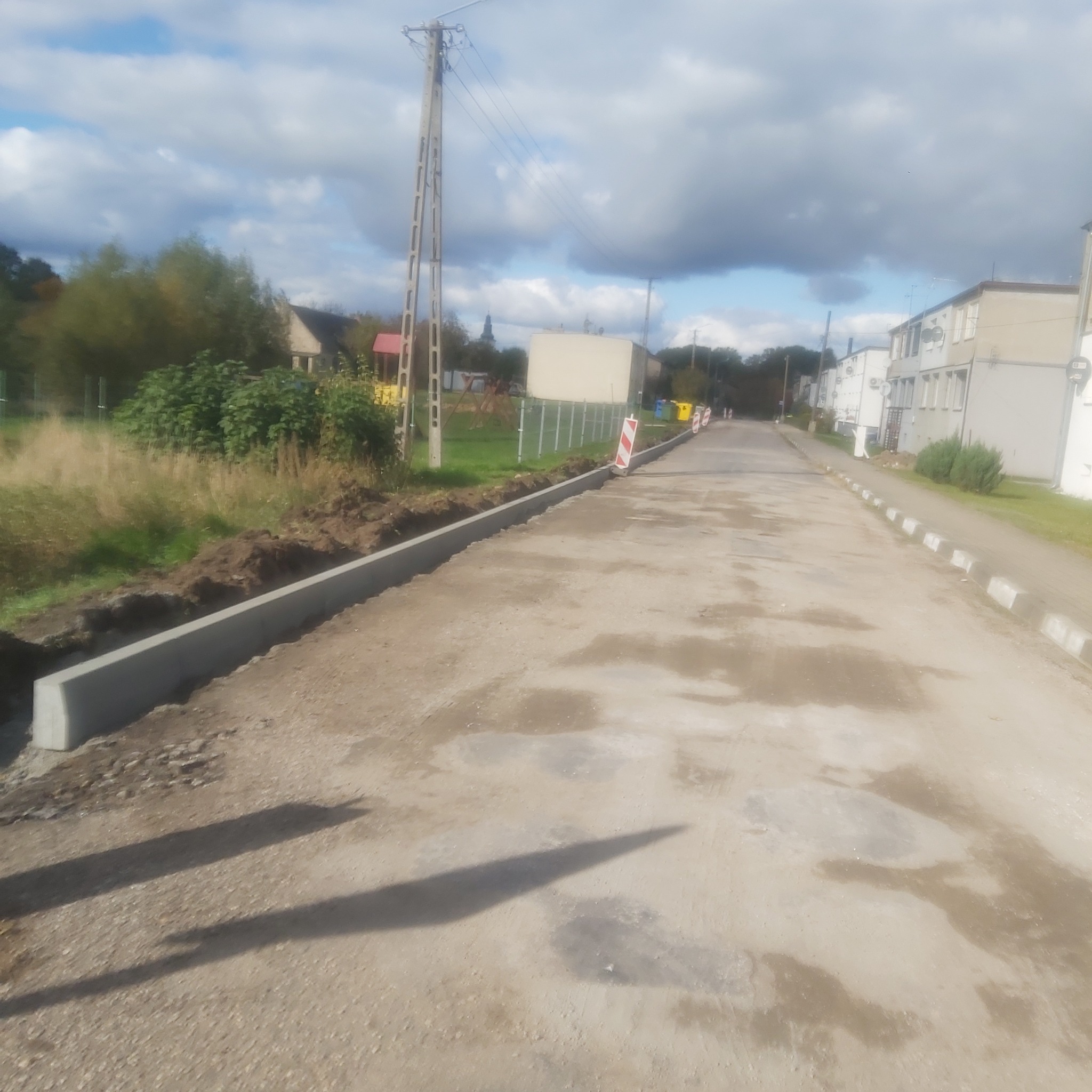 Świeżo wylany betonowy obrzeżnik chodnika wzdłuż drogi w trakcie budowy, z widocznymi znakami ostrzegawczymi i elementami krajobrazu w tle.