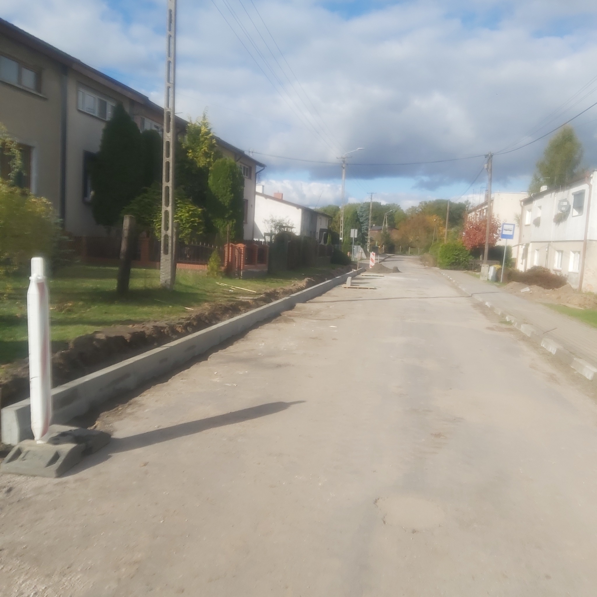 Betonowe obrzeża chodnika w trakcie budowy, widoczny słupek geodezyjny i przygotowane podłoże w tle zabudowa wiejska.