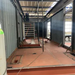 Wnętrze kontenera modułowego w trakcie budowy, widoczne stalowe ramy, podłoga z blachy ryflowanej, schody na rusztowaniu oraz pracownik na górze.