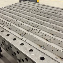 Stół spawalniczy z zamocowanymi i wstępnie pospawanymi elementami konstrukcyjnymi, w tle wózek widłowy i wyposażenie warsztatu.