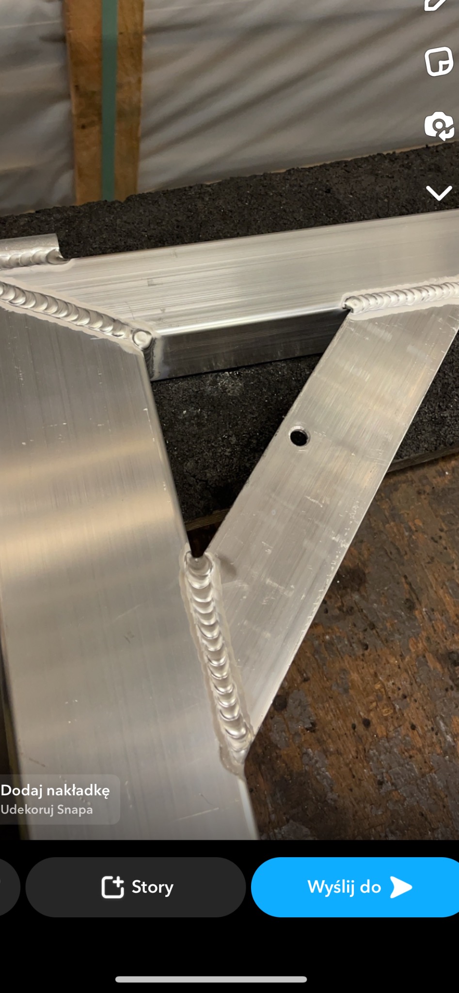 Fragment konstrukcji aluminiowej z widocznymi spawami, wykonany z profili o przekroju kwadratowym, z otworem montażowym, leżący na ciemnym podłożu.