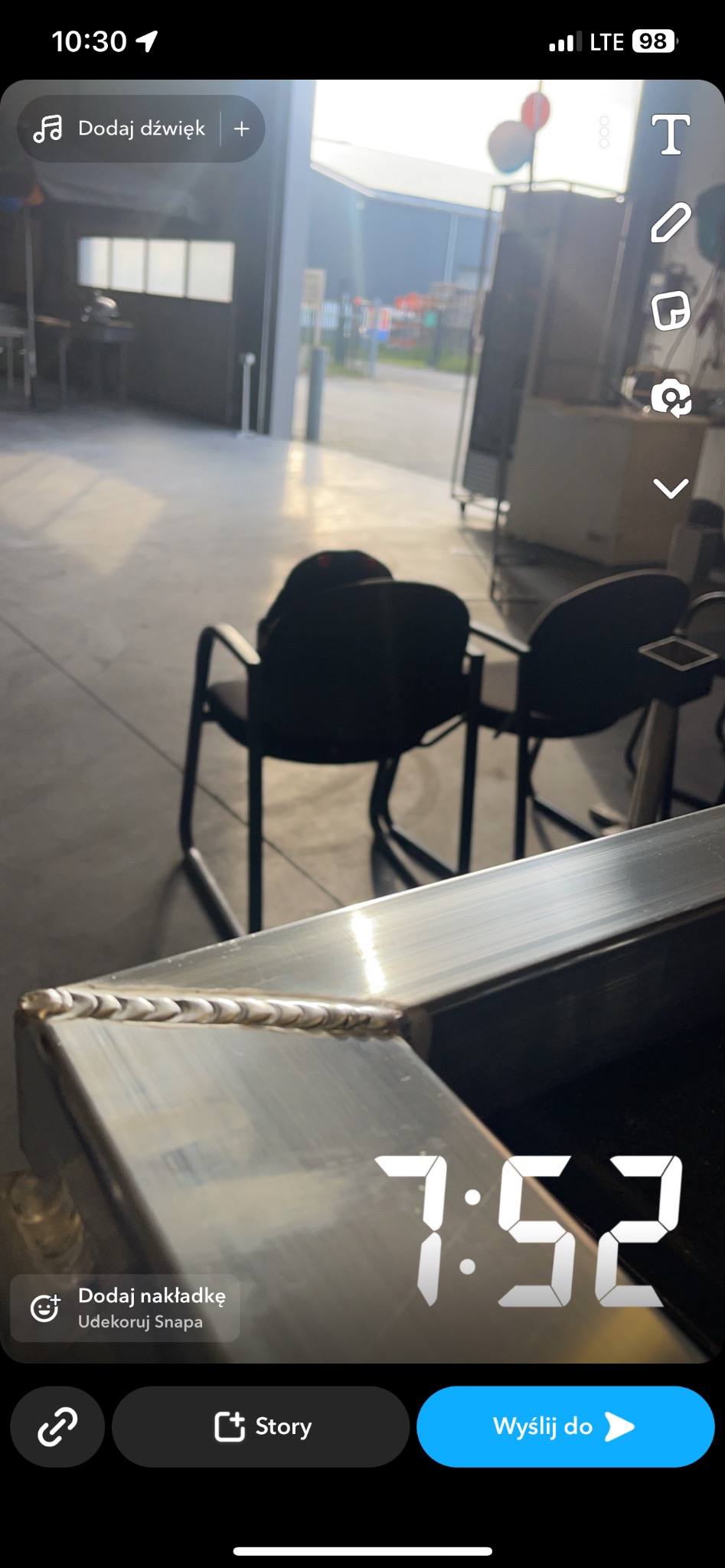 Spoina na metalowym profilu w warsztacie, widoczne krzesła i brama w tle, zdjęcie zrobione telefonem z nakładkami aplikacji.