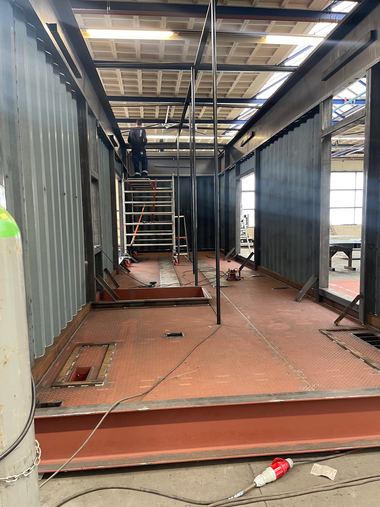 Wnętrze kontenera modułowego w trakcie budowy, widoczne stalowe ramy, podłoga z blachy ryflowanej, schody na rusztowaniu oraz pracownik na górze.