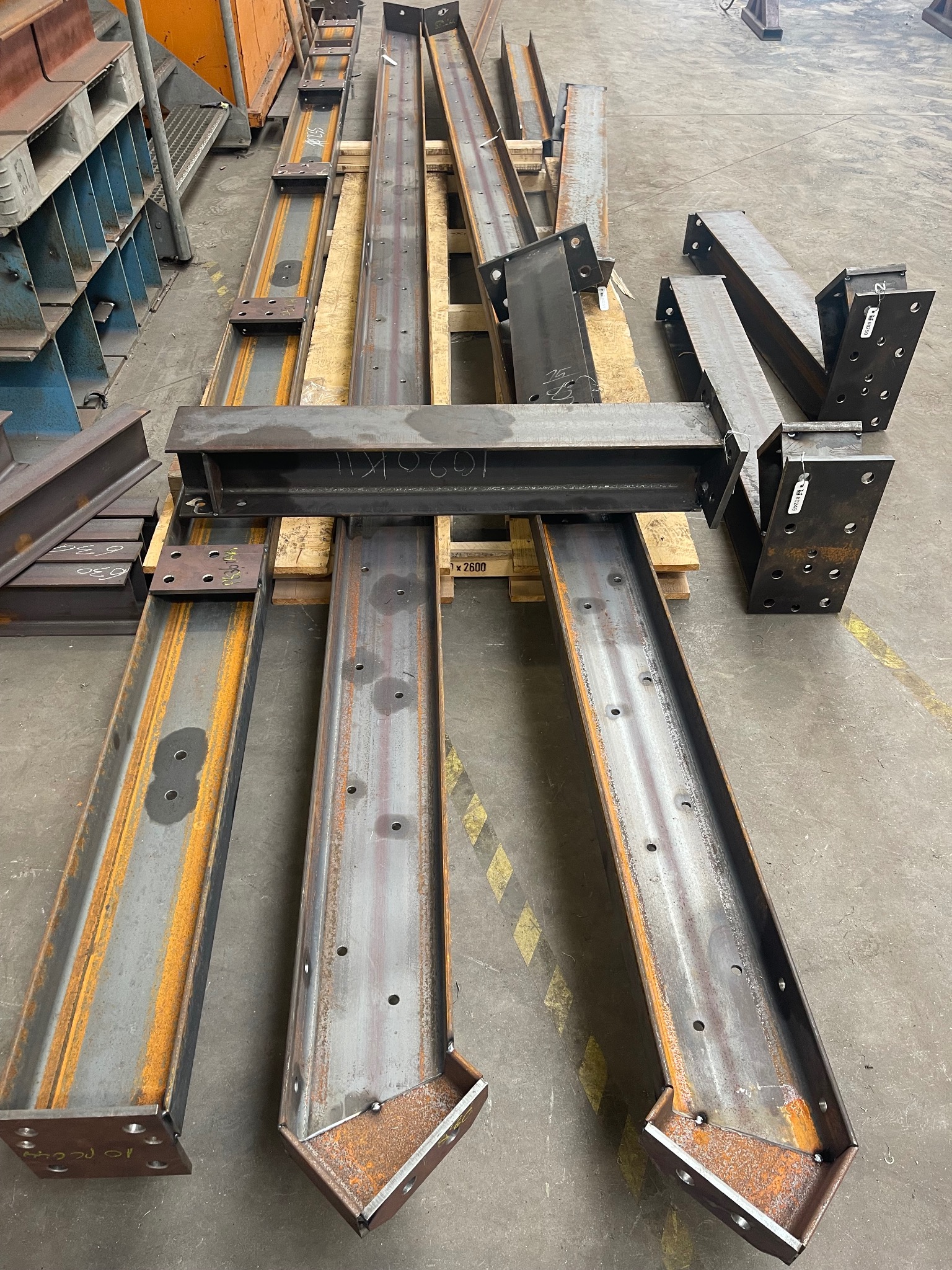 Przygotowane elementy konstrukcyjne ze stali, profile dwuteowe z nawierconymi otworami, ułożone na drewnianych paletach w hali produkcyjnej, widoczne ślady rdzy i spawania.