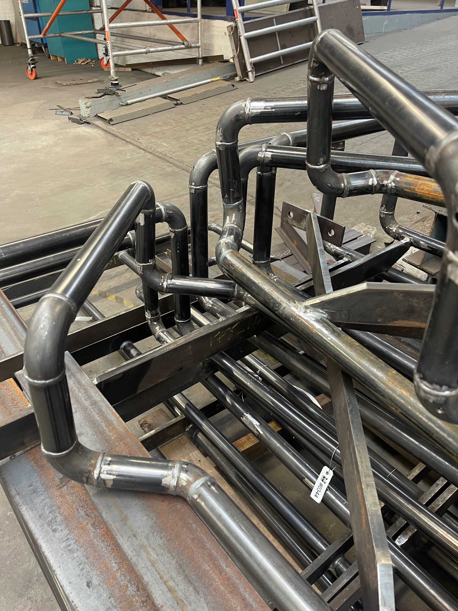 Stos spawanych, metalowych rur i profili o różnych kształtach, w warsztacie, z widocznymi śladami spawania i etykietą identyfikacyjną.