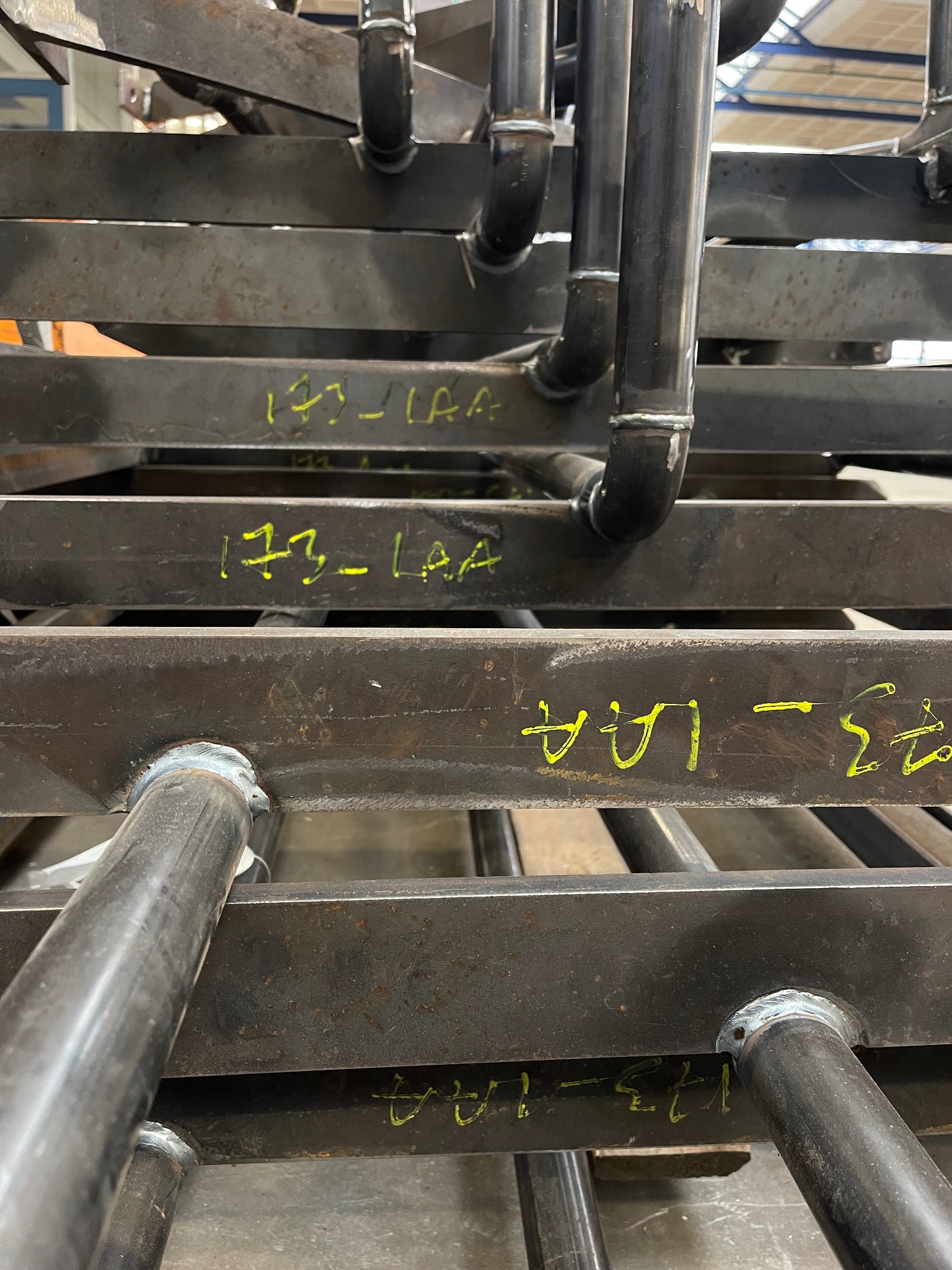 Metalowa konstrukcja z rur i profili prostokątnych połączonych spawami, z żółtymi oznaczeniami naniesionymi na powierzchnię.