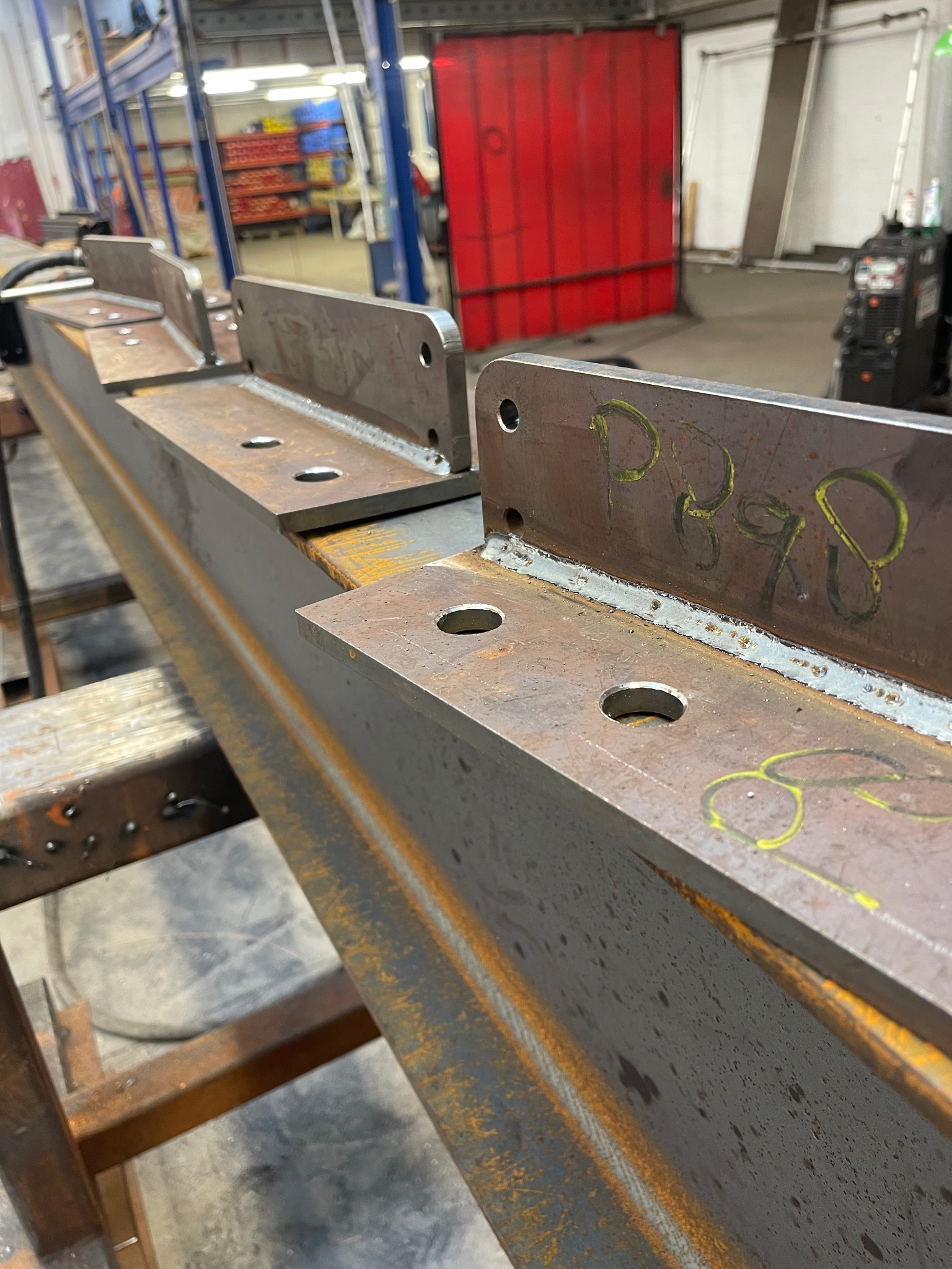 Metalowa belka z przyspawanymi elementami mocującymi, otwory na śruby, oznaczenia żółtą farbą, widoczne spawy, konstrukcja wsporcza w tle, hala produkcyjna.