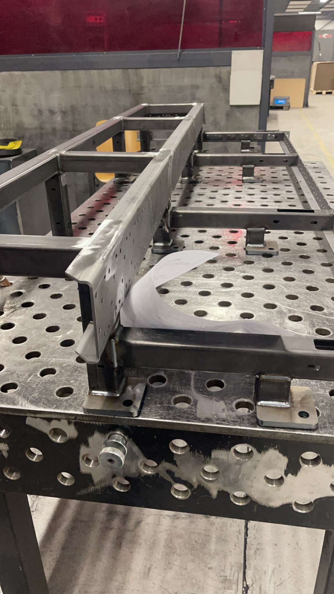 Metalowa konstrukcja ramowa z otworami, częściowo spawana, leżąca na perforowanym stole warsztatowym, obok planów technicznych.