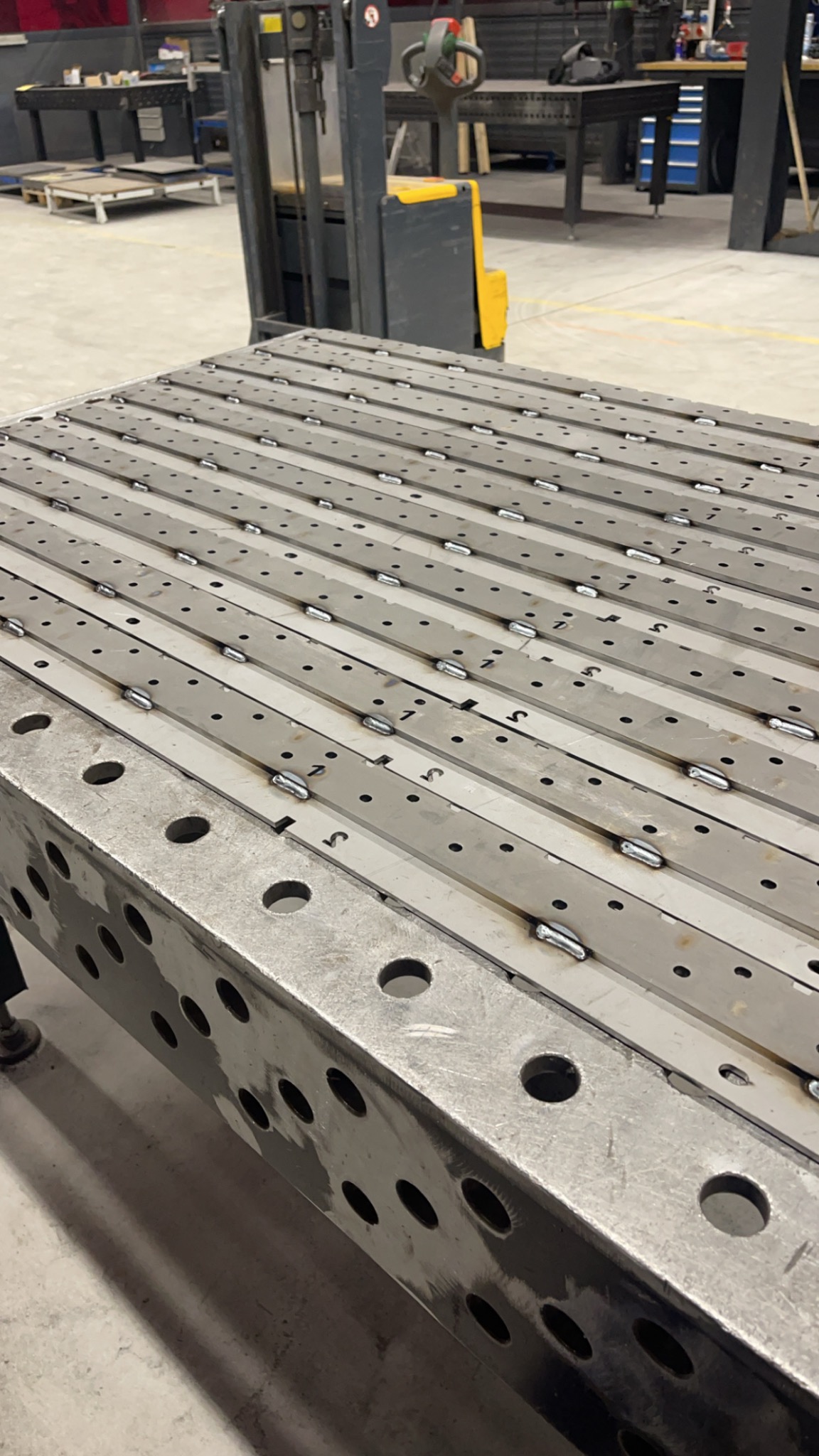 Stół spawalniczy z zamocowanymi i wstępnie pospawanymi elementami konstrukcyjnymi, w tle wózek widłowy i wyposażenie warsztatu.