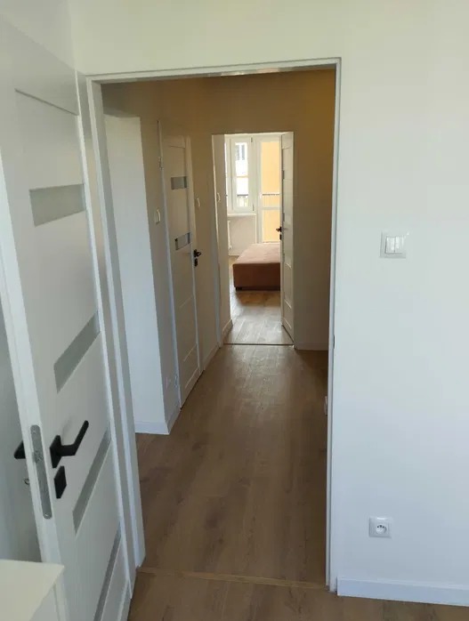 Jasny korytarz z drewnianą podłogą i białymi drzwiami. Widok w głąb mieszkania, z oknem w tle. Minimalistyczny design.